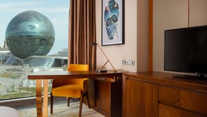 Habitación de hotel con escritorio, silla, televisión y un amplio ventanal con vistas a un edificio moderno.