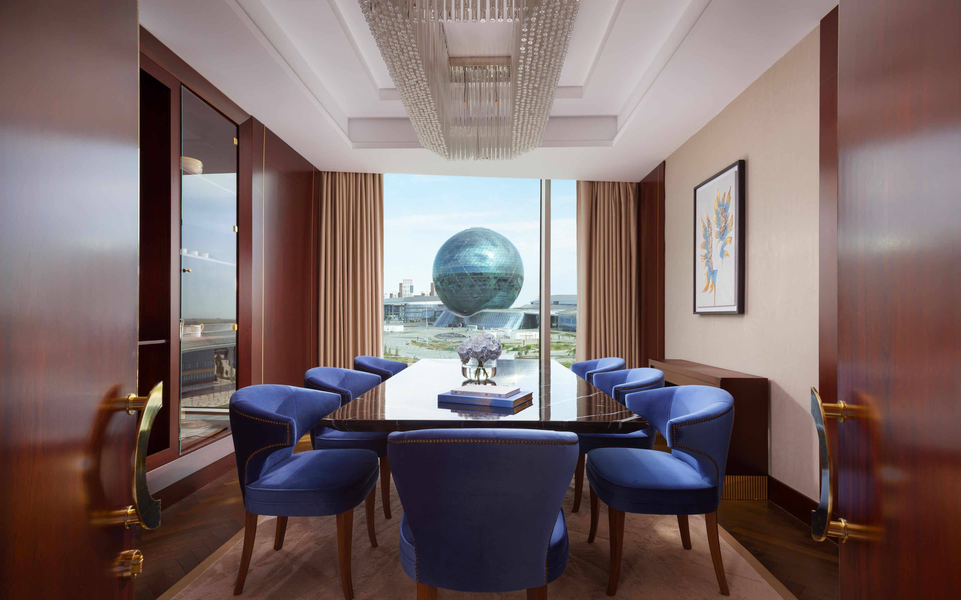 A conference room with a large table, blue chairs, a chandelier, a framed artwork, and a view of a modern building with a spherical structure.