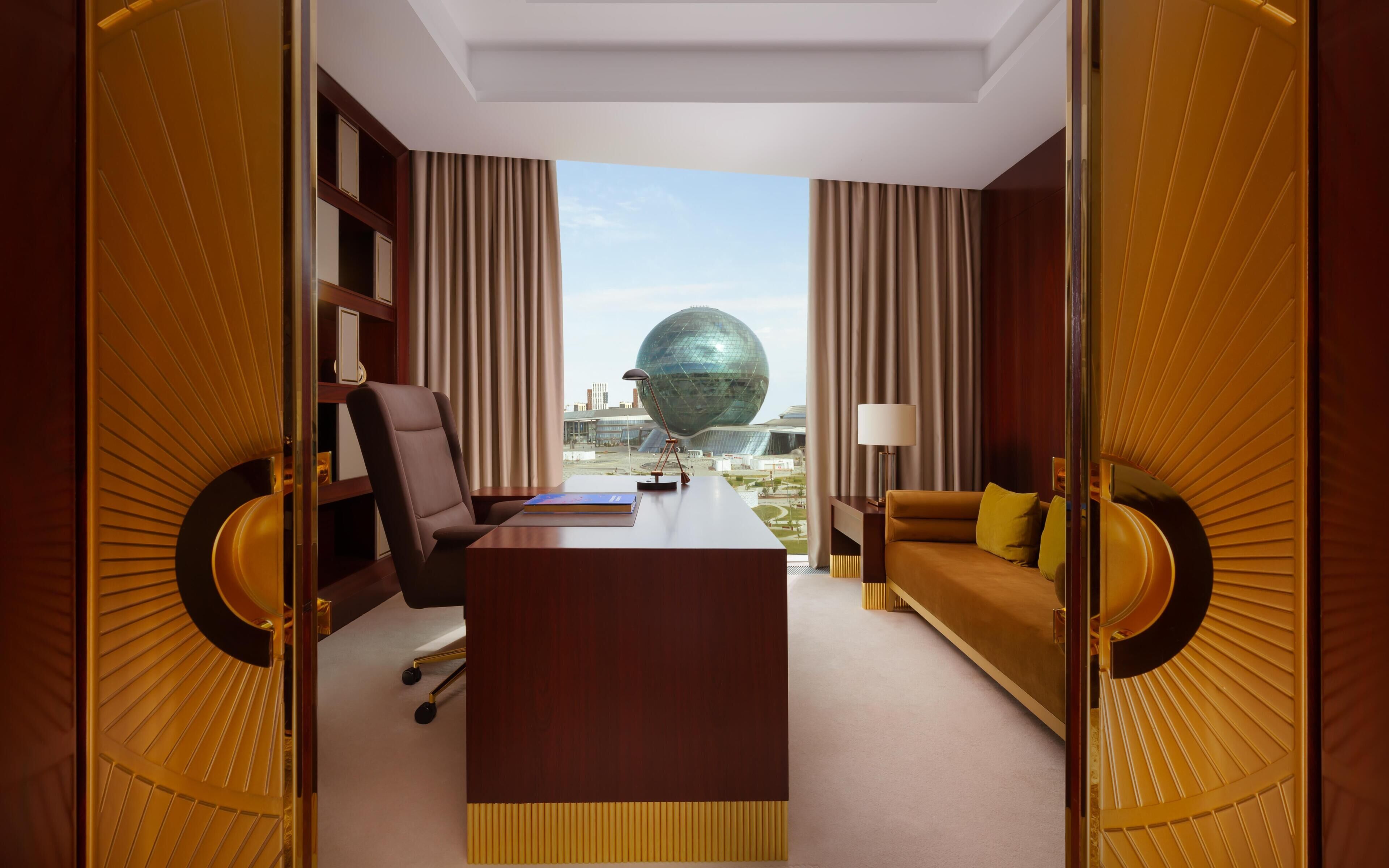 A hotel room with a desk, chair, sofa, and a view of a modern building.