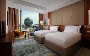 Habitación de hotel con dos camas, un escritorio, una silla, un televisor y vistas a un edificio moderno.