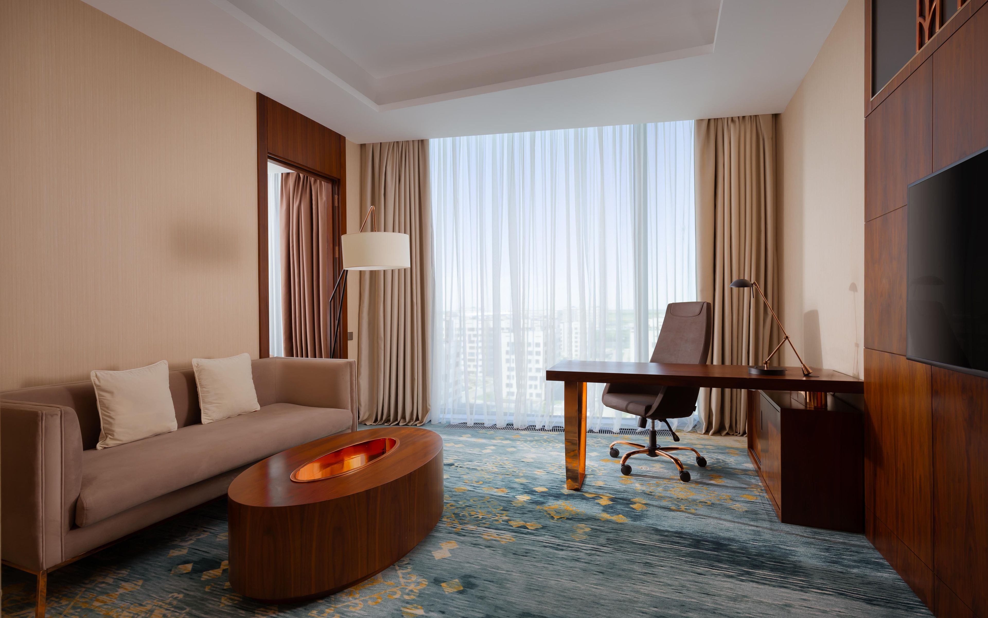 Habitación de hotel con sofá, escritorio, silla, televisión y un gran ventanal con cortinas.