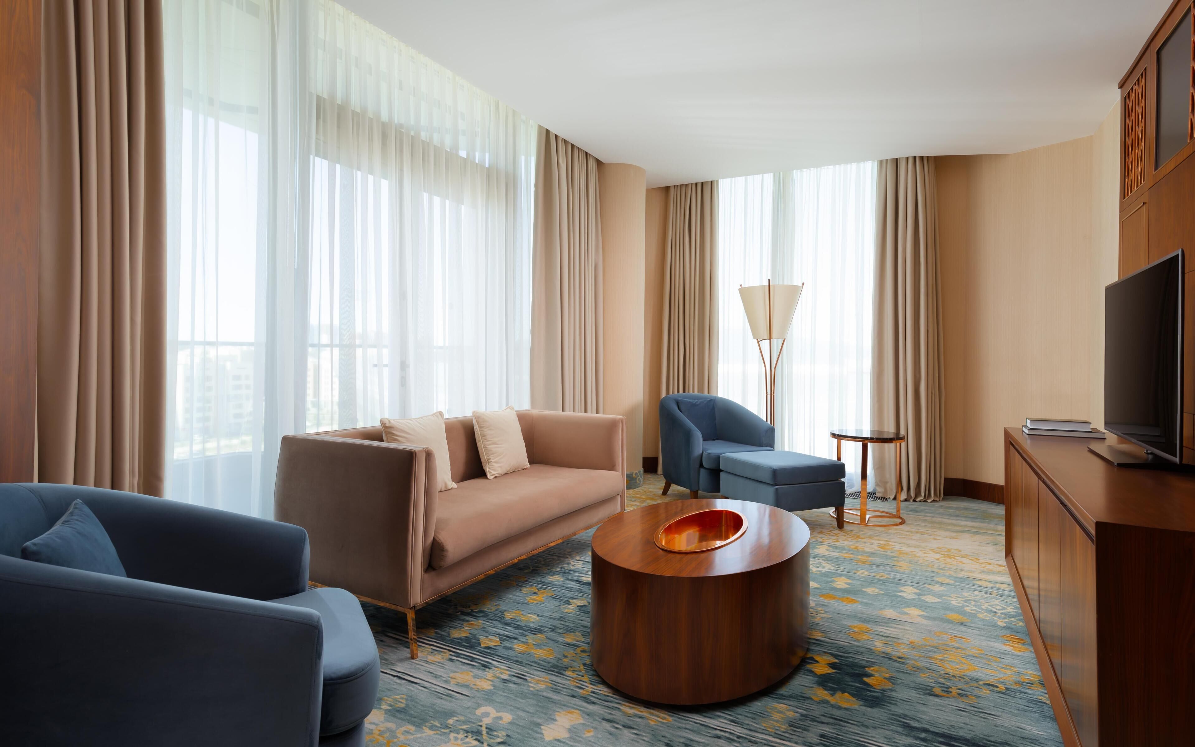A modern hotel room with a sofa, armchairs, a coffee table, a TV, and a lamp.