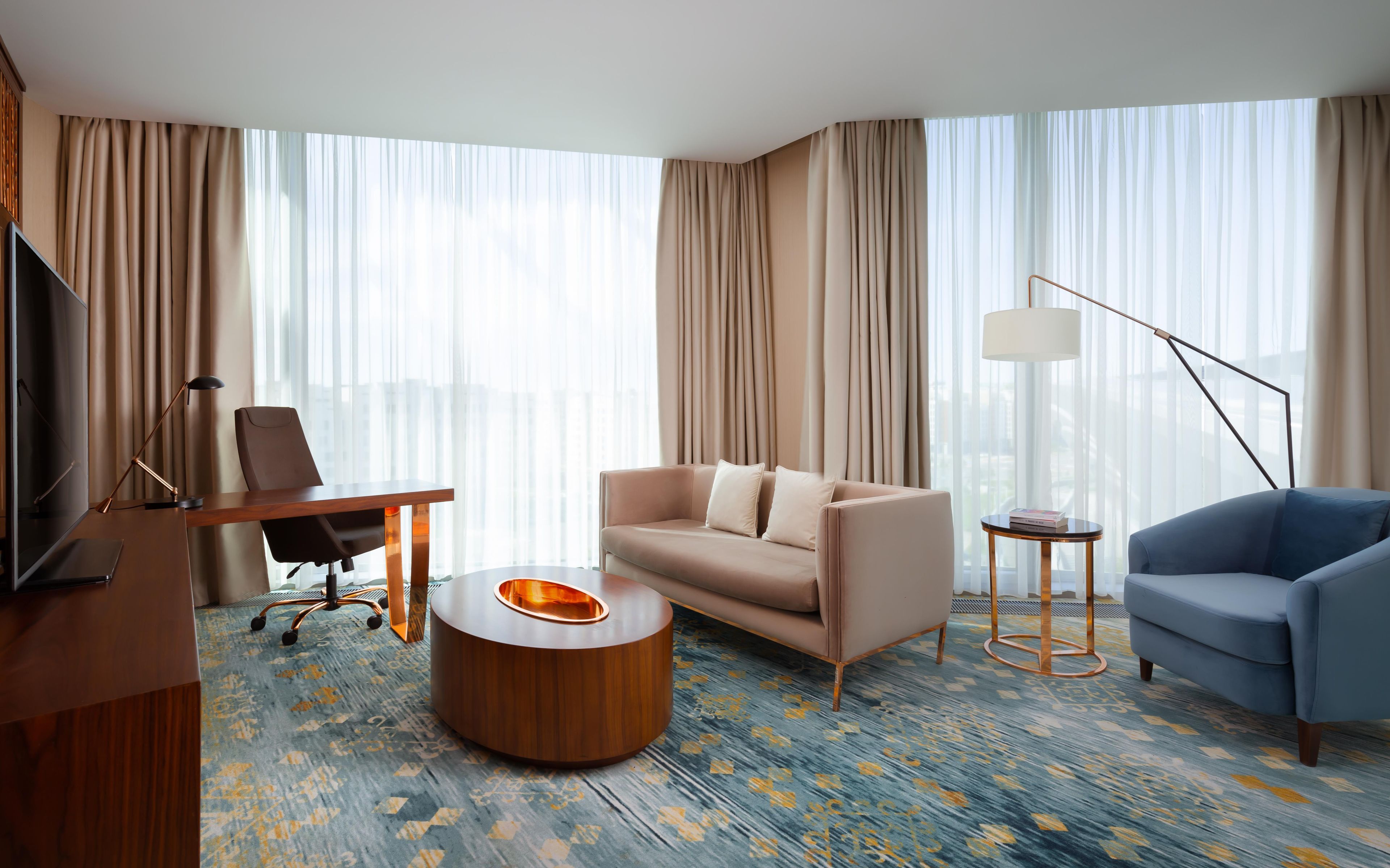 A modern hotel room with a sofa, a blue armchair, a wooden coffee table, a desk with a chair, a TV, and large windows with curtains.