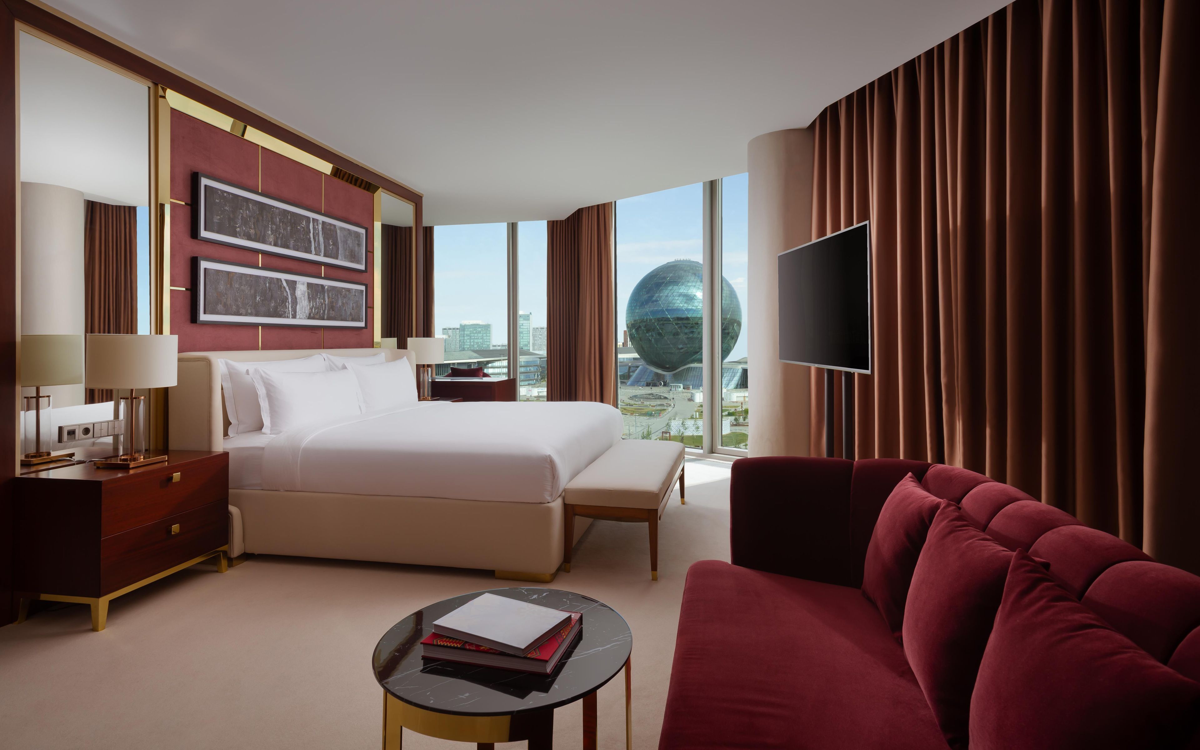 Habitación de hotel con una cama grande, televisor, sofá rojo, una mesita con libros y vistas a la ciudad.