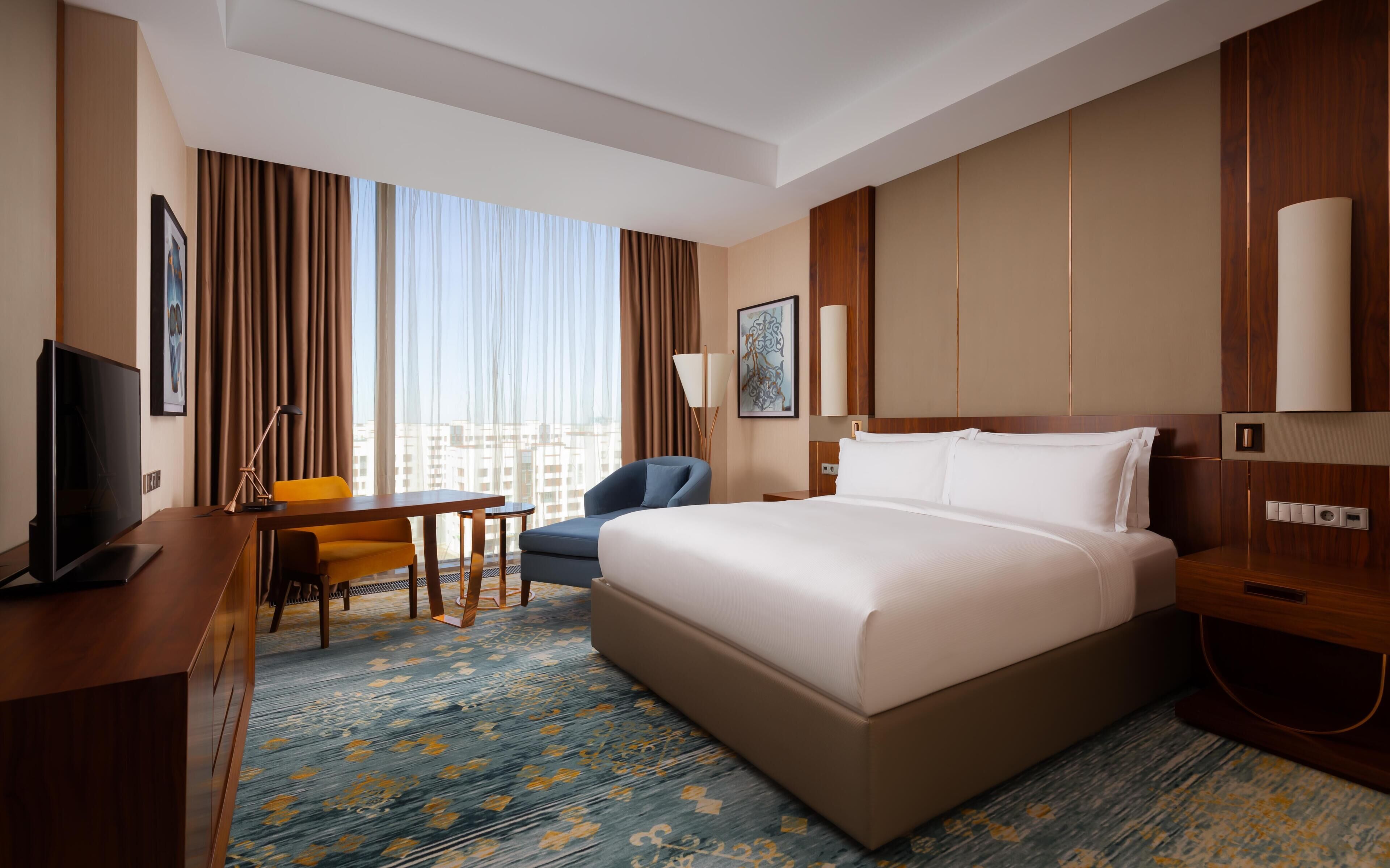 Una habitación de hotel con una cama, un escritorio con televisor, una silla, una lámpara y vistas a la ciudad.
