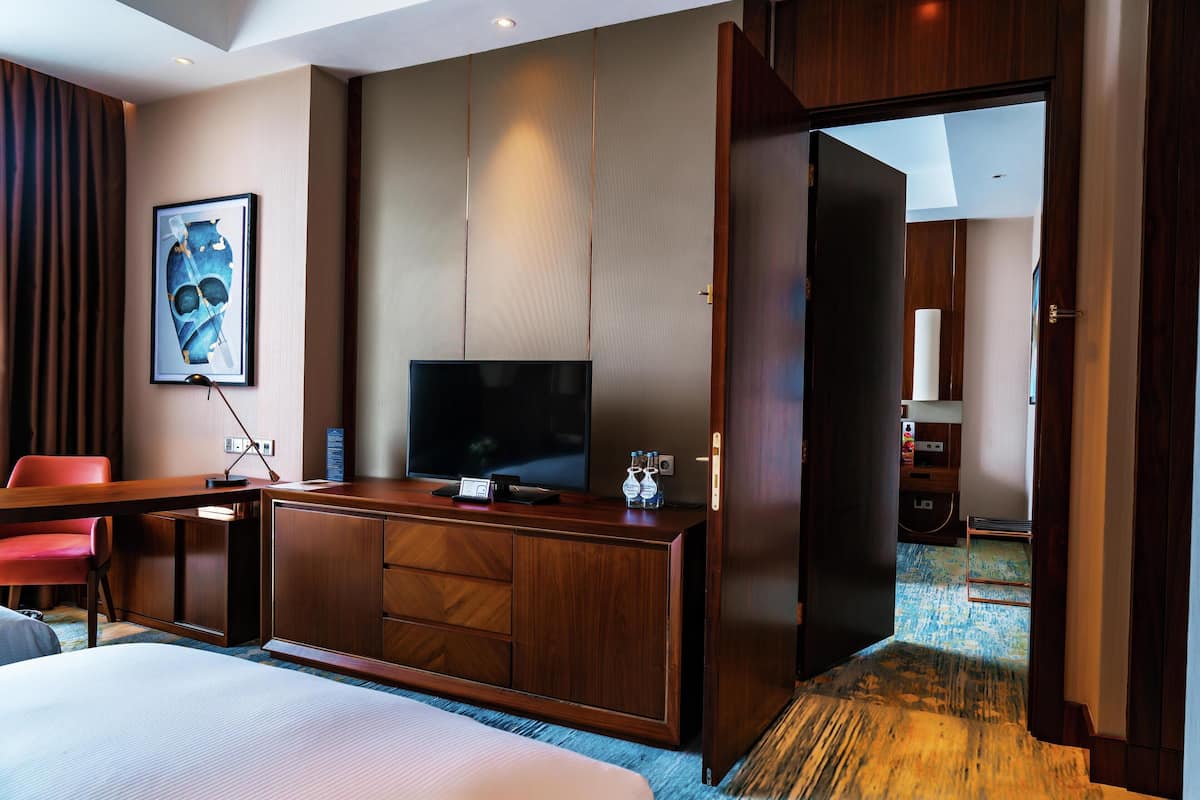Habitación de hotel con una cama, un escritorio con televisor, una silla roja, un armario de madera y una puerta que da a otra habitación.