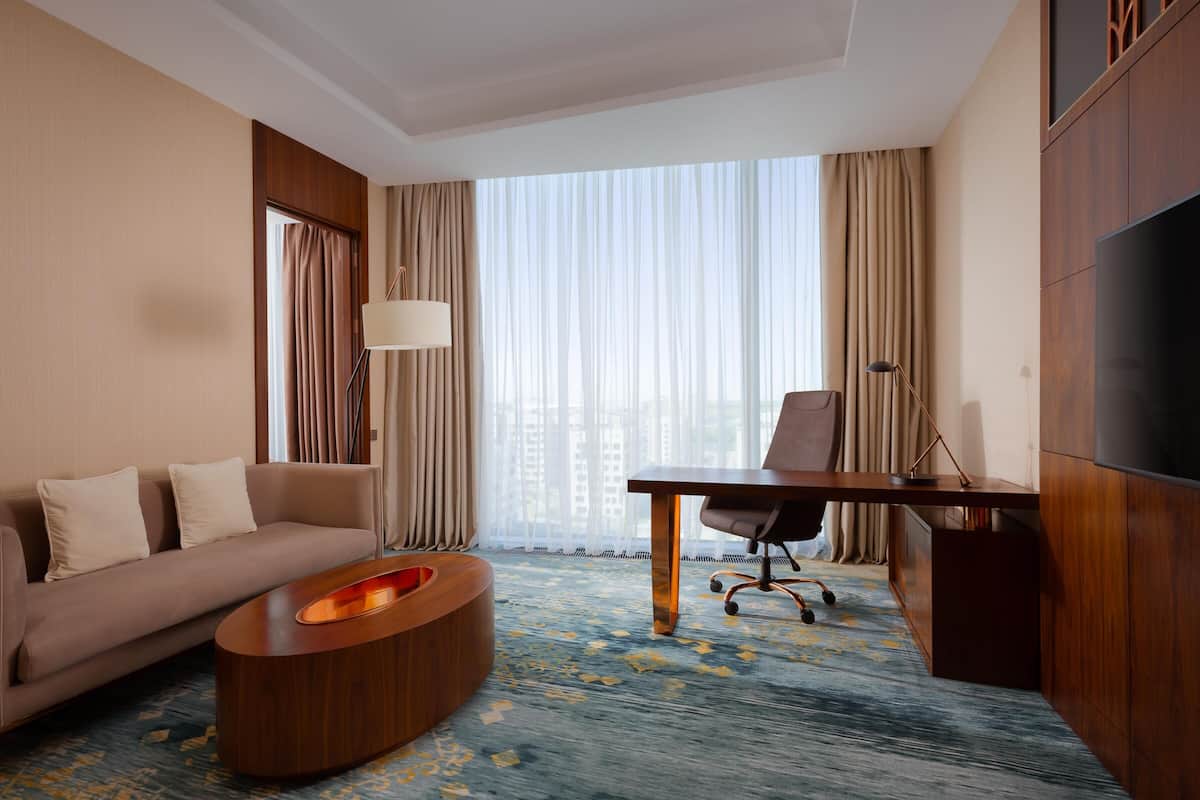 Habitación de hotel con sofá, escritorio, silla, televisión y un gran ventanal con cortinas.