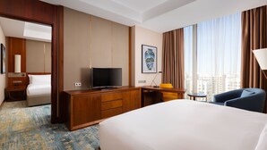 Habitación de hotel con una cama grande, un escritorio con televisor, una silla y vistas de la ciudad a través de grandes ventanales.
