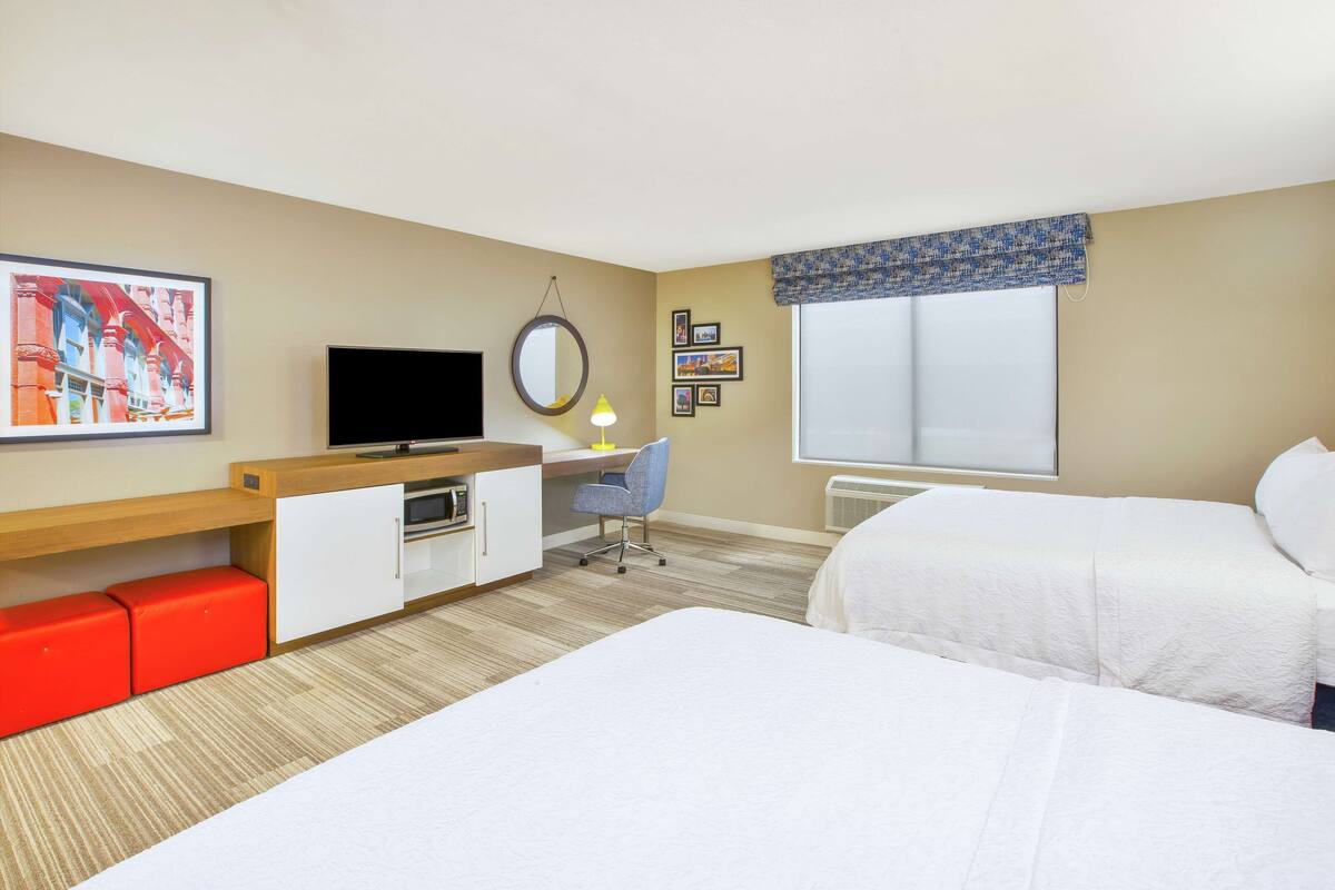 A hotel room with two beds, a desk with a chair, a television, and a window with a patterned valance.