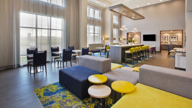 Um lobby de hotel moderno com áreas de estar, um bar e uma cozinha visíveis ao fundo.