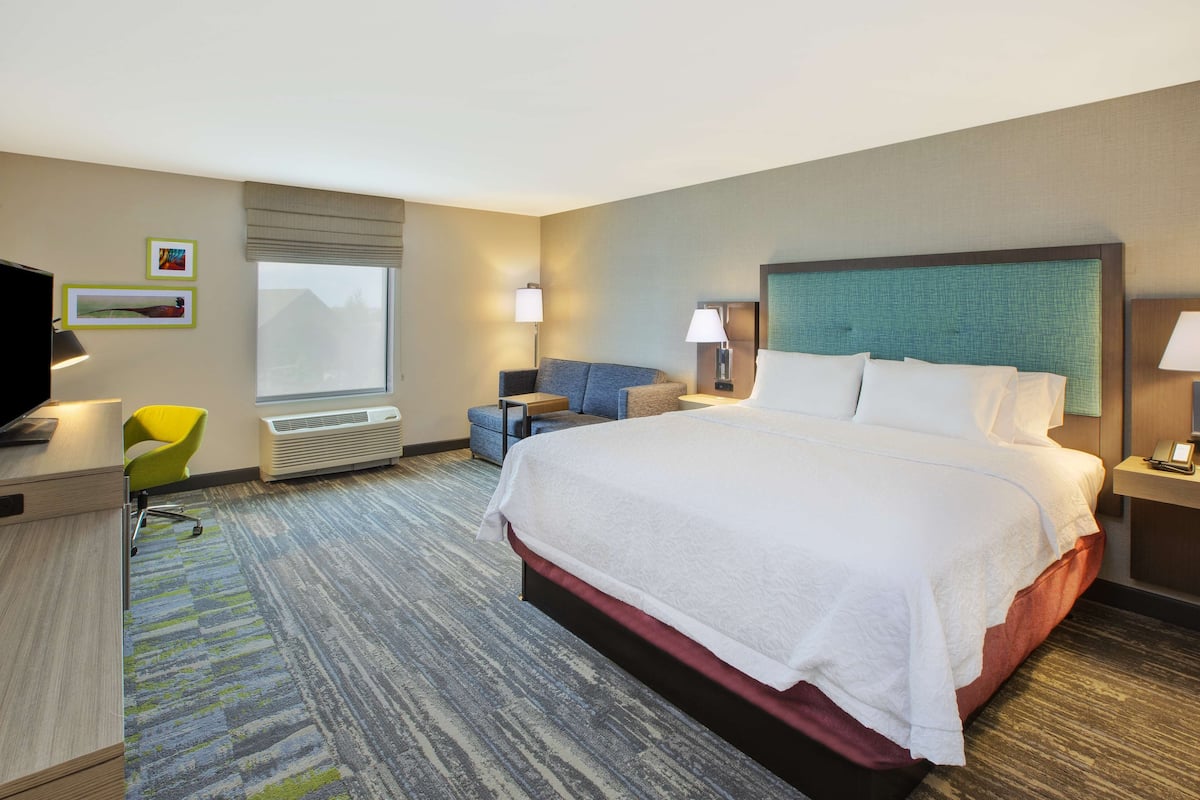A hotel room with a large bed, a desk with a chair, a TV, and a window with blinds.