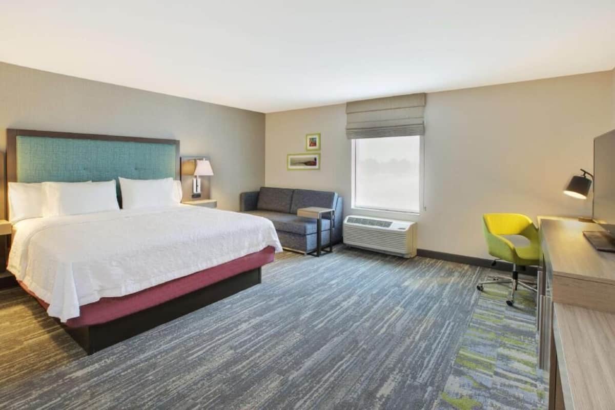 A hotel room with a bed, a sofa, a desk, a chair, a lamp, and a window.