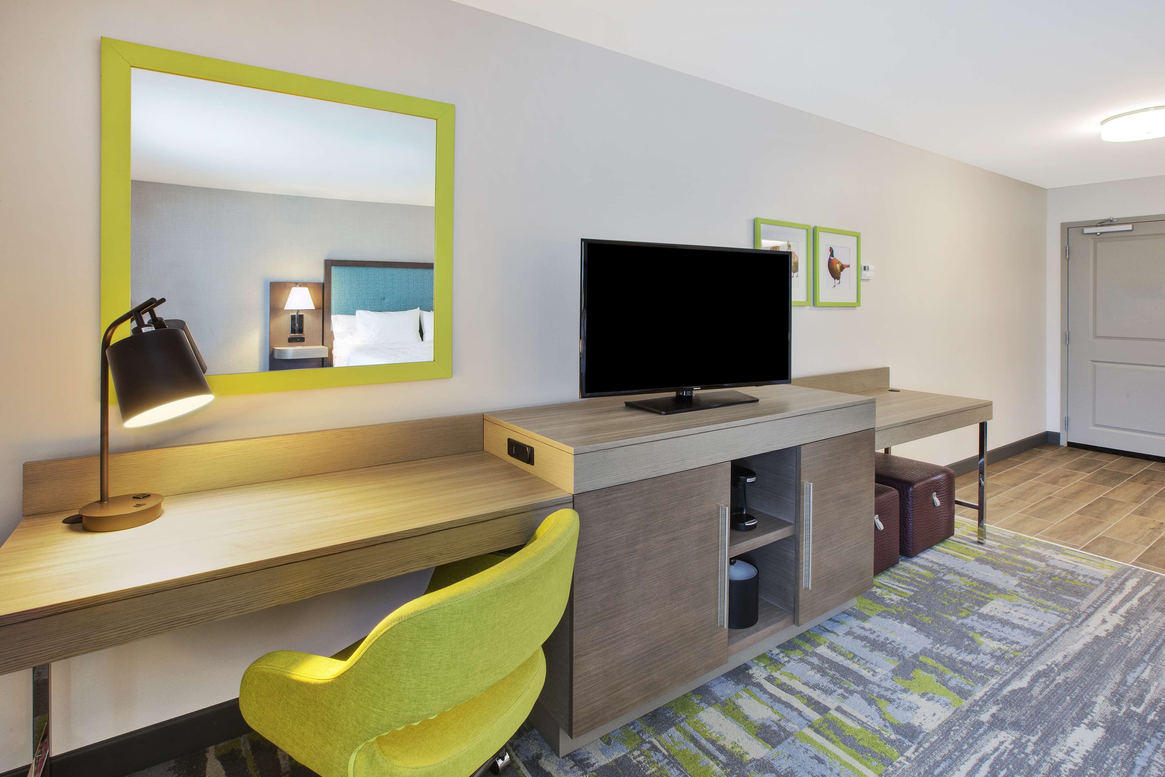 A hotel room with a desk, a television, a mirror, a chair, and a bed.