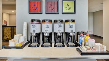 A coffee station with four dispensers, each labeled with a different type of coffee. There are stacks of disposable cups and a tray with napkins and condiments.