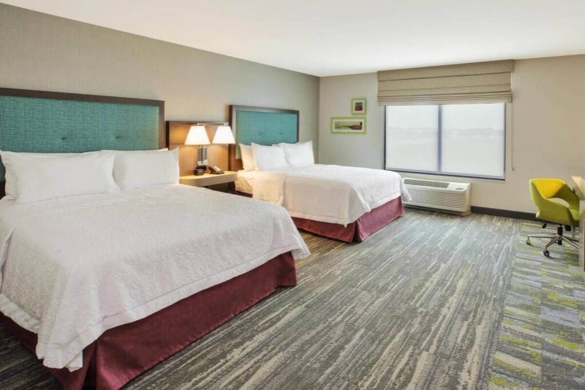 A hotel room with two beds, a nightstand with a lamp, a chair, and a large window.