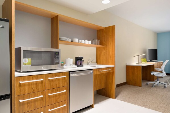 Una cocina moderna con microondas, cafetera y lavavajillas. Hay un estante de madera sobre la encimera y un escritorio con una silla al fondo.