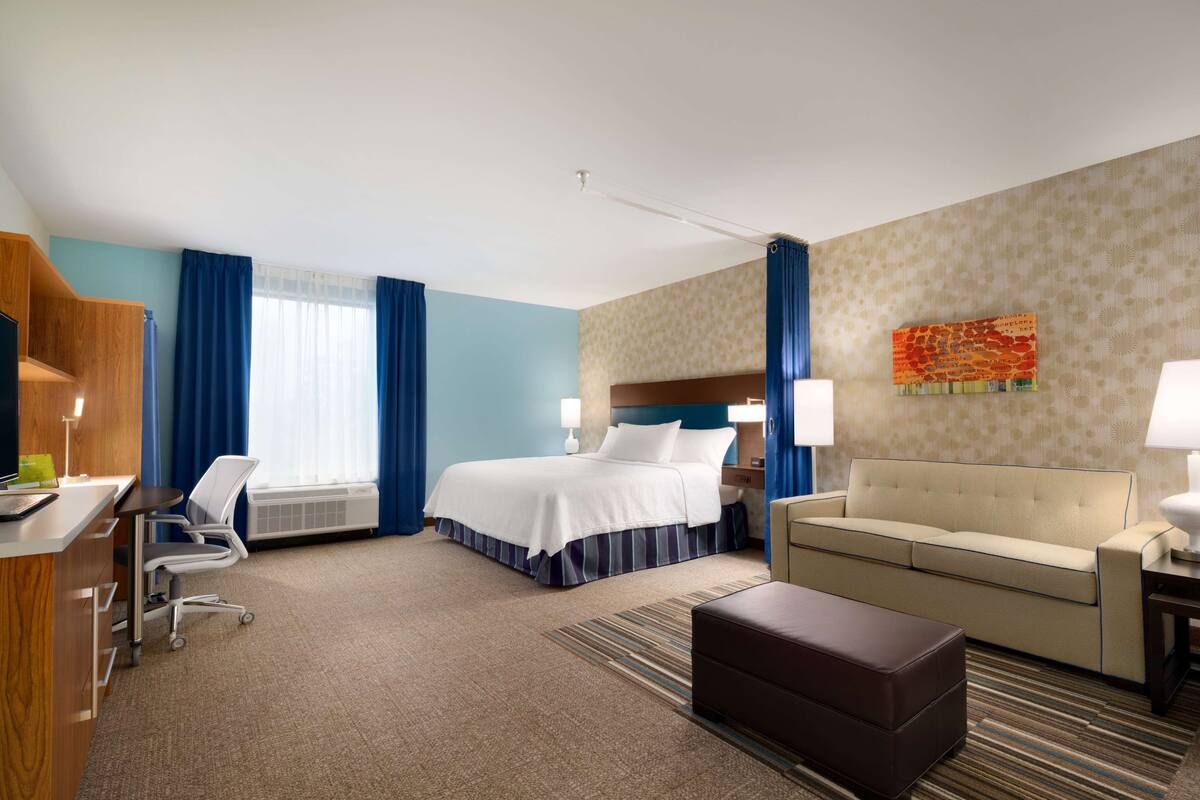 Une chambre d’hôtel avec un lit, un bureau, une chaise, un canapé et une grande fenêtre avec des rideaux.