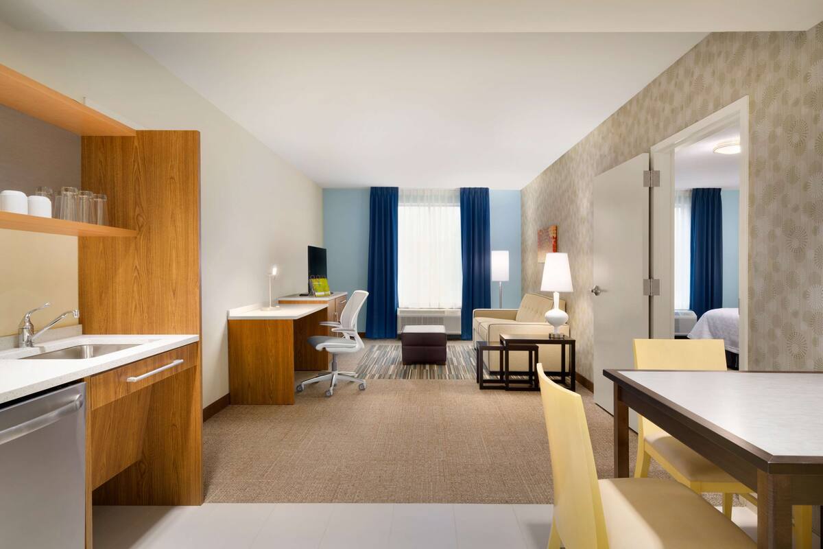 Une chambre d’hôtel équipée d’une kitchenette, d’un bureau avec une chaise, d’une table à manger avec des chaises, d’un canapé et d’un grand miroir.