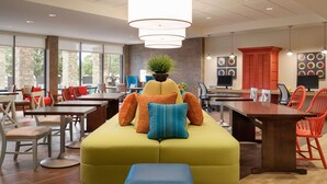 A modern dining area with a large wooden table, multiple chairs, and a yellow sofa with orange and blue cushions.