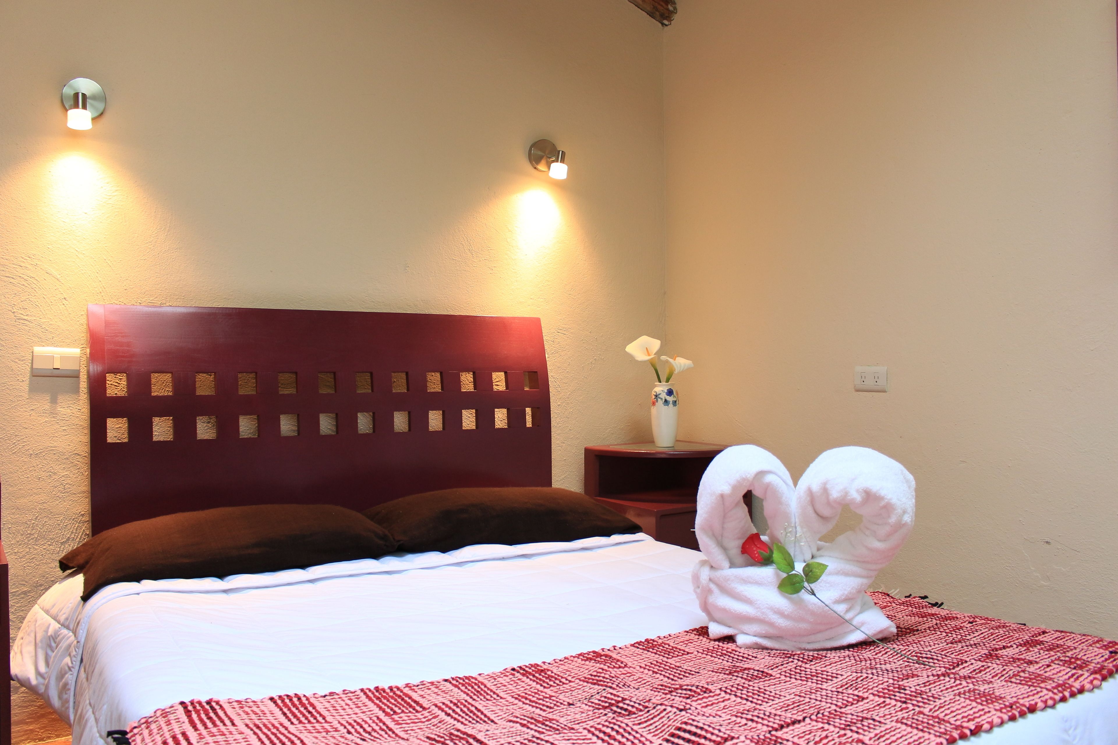 A hotel room with a bed, a headboard, a nightstand, a vase with a flower, and two towels shaped like swans.