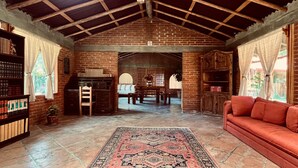 Una habitación amplia con techo alto, vigas de madera a la vista, chimenea de ladrillo, alfombra grande, sofá y estantería.
