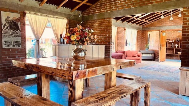 Un interior rústico con una mesa de madera, bancos, un sofá rosa y una pared de ladrillo.