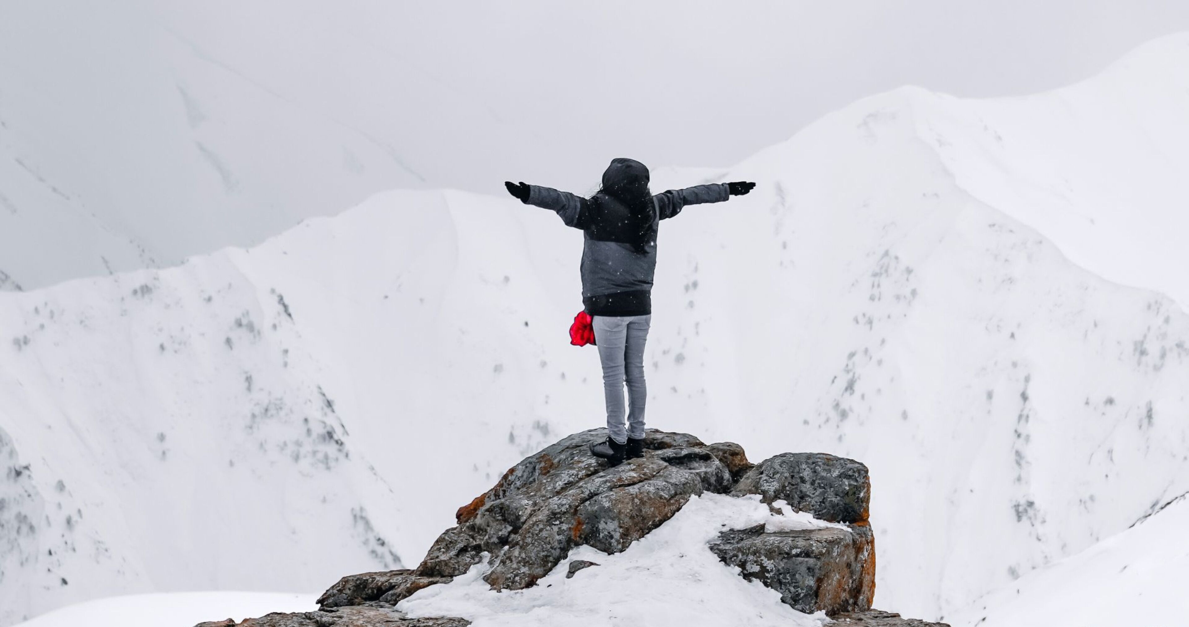Una persona de pie sobre una roca con los brazos extendidos, rodeada de montañas cubiertas de nieve.