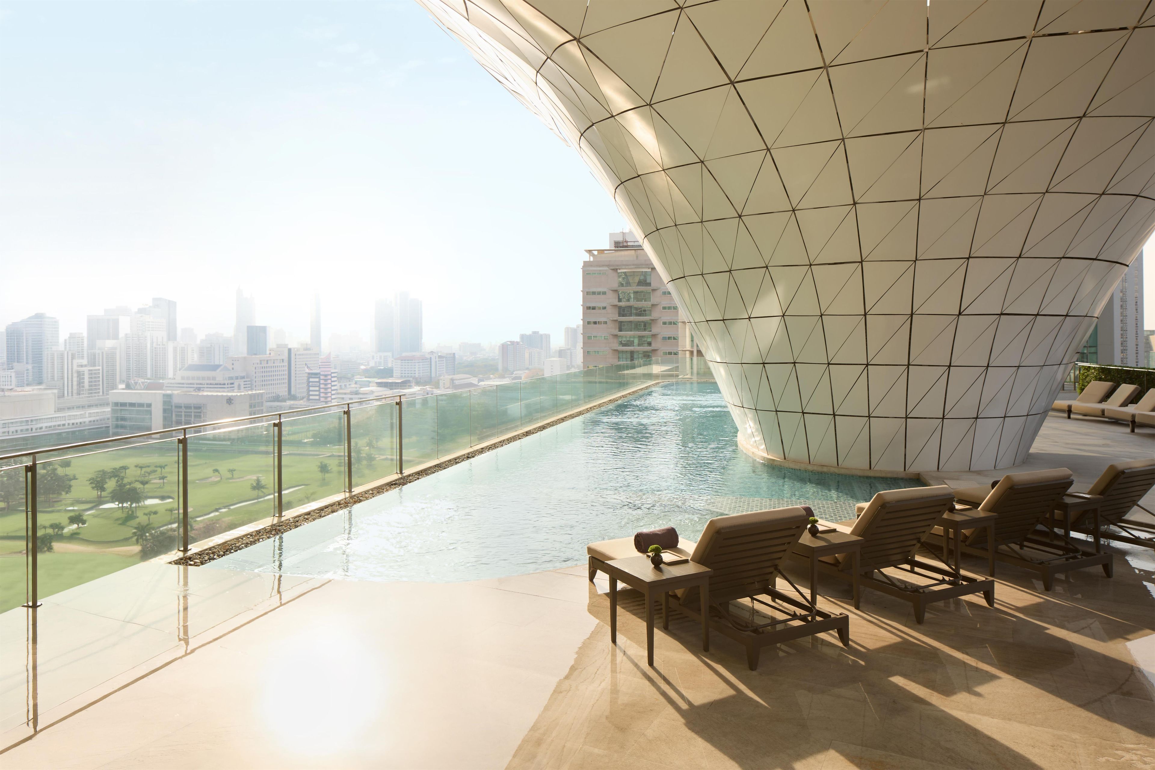 A modern building with a unique curved roof design, overlooking a swimming pool and a cityscape.