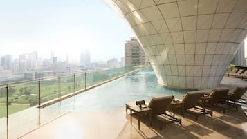 A modern building with a unique curved roof design, overlooking a swimming pool and a cityscape.