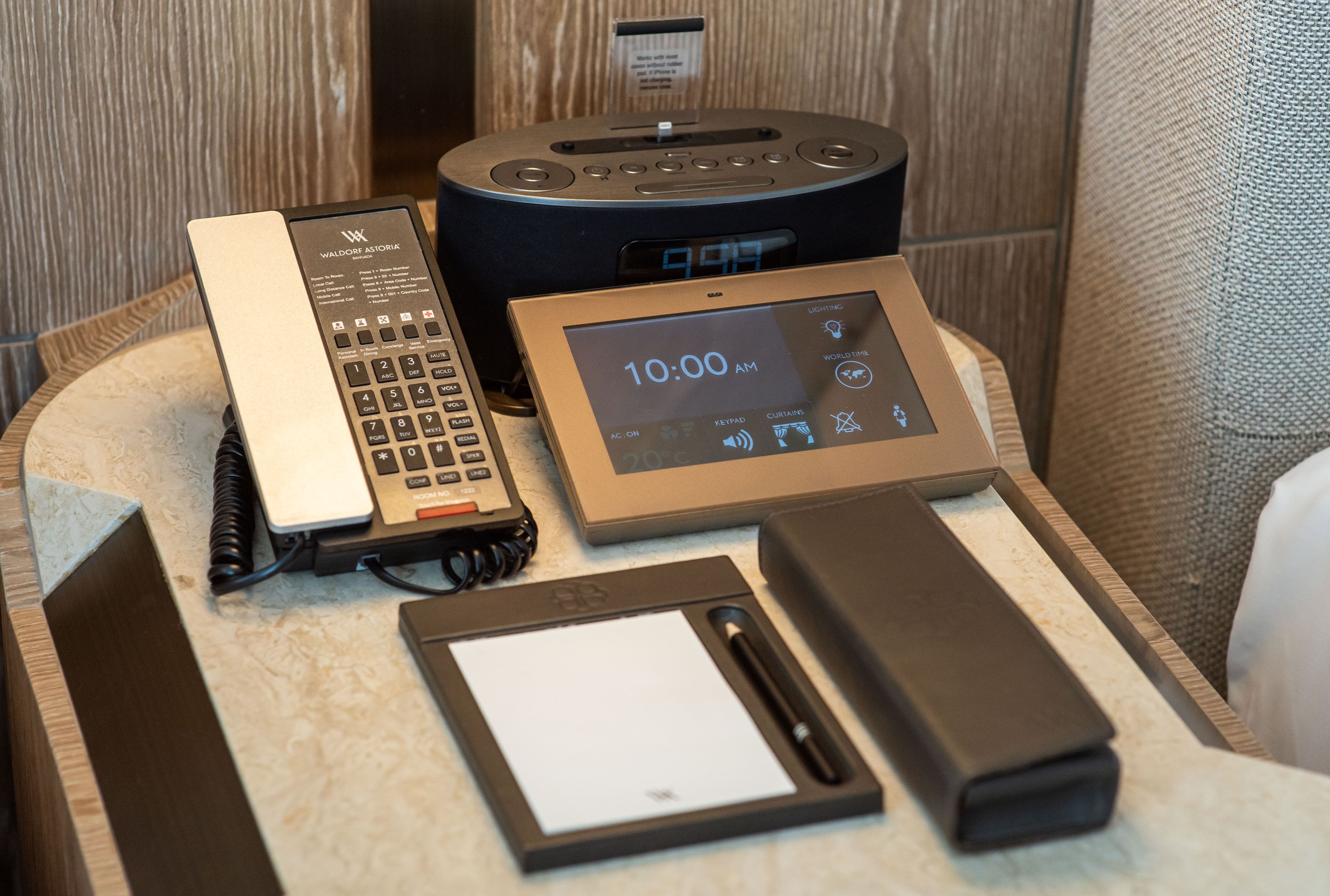 Ein Hotelrezeptionstresen mit einer digitalen Uhr, die 10:00 Uhr anzeigt, einem Festnetztelefon, einem Tablet, einem Stift und einer Ladestation.