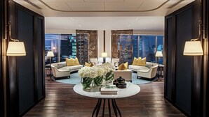 Een moderne hotellobby met donker houten vloeren, een ronde tafel met een boek en vazen, twee banken met gele kussens, en grote ramen met uitzicht over de stad.