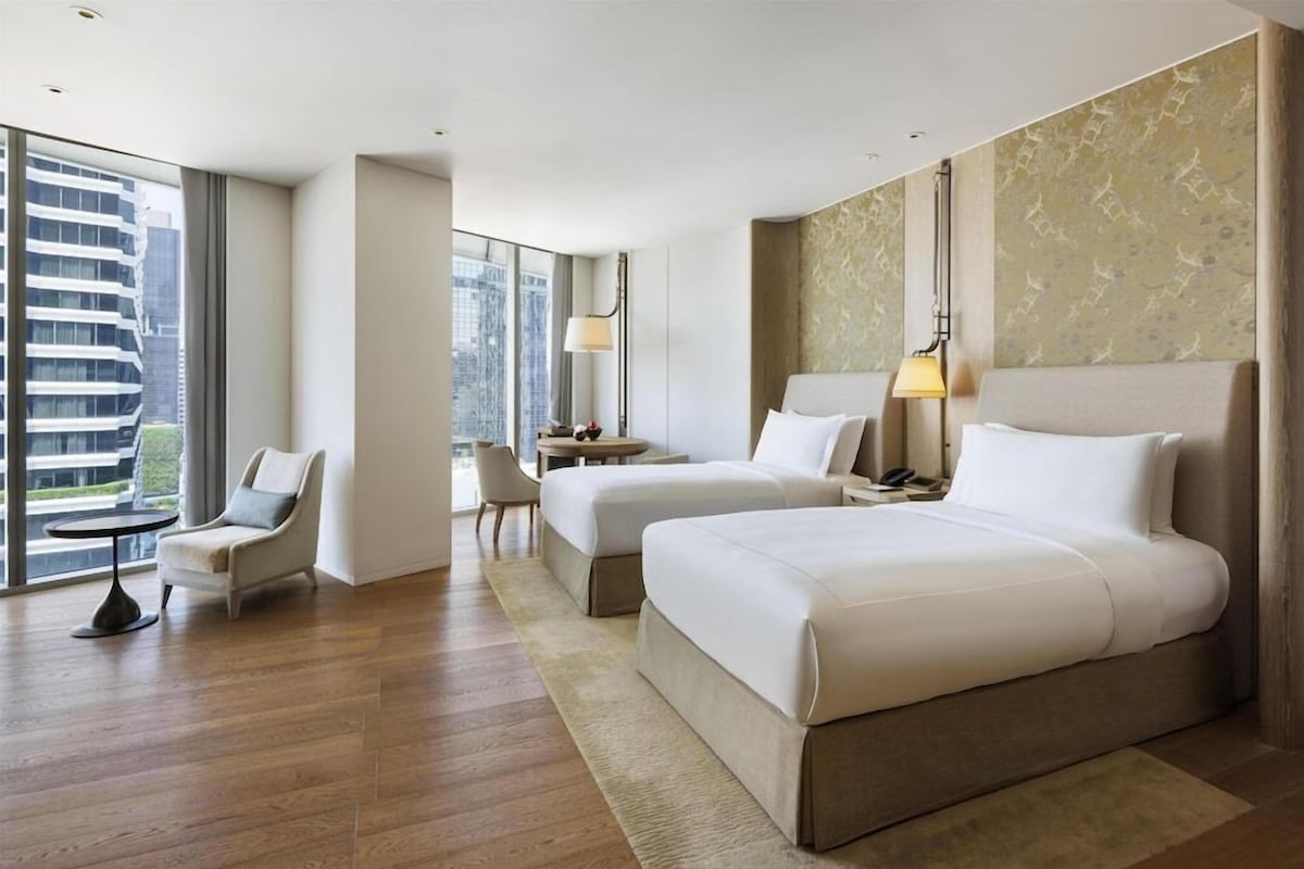 A hotel room with two beds, a sitting area with a chair and table, a large window with a city view, and a patterned wall.