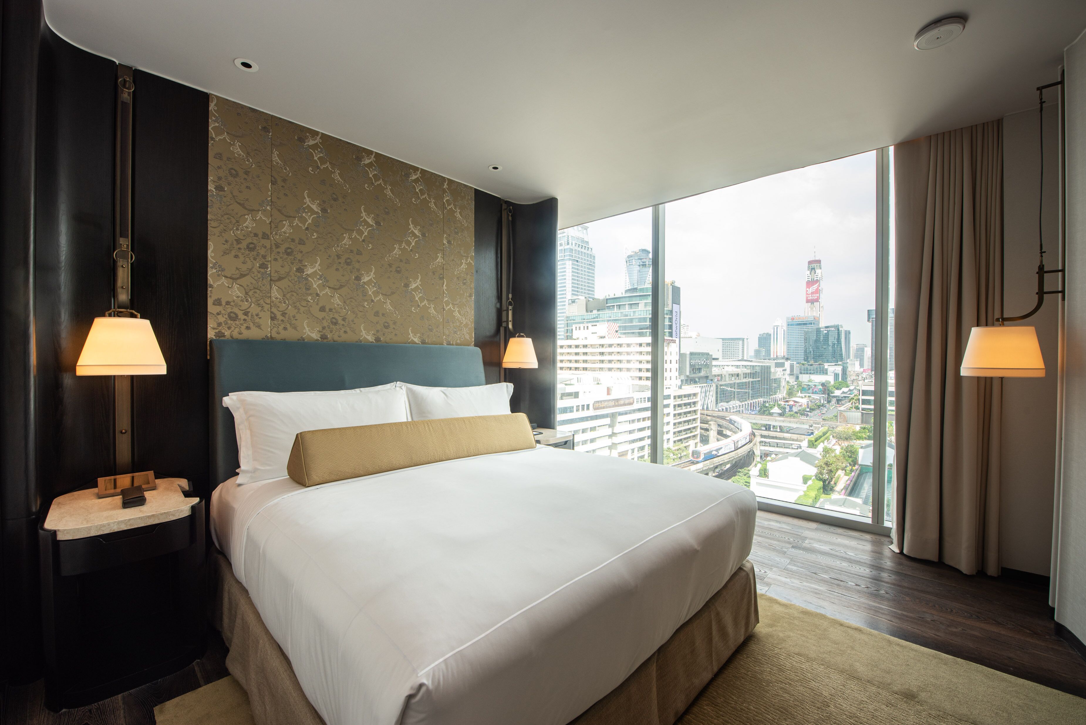 A modern hotel room with a large bed, two bedside tables with lamps, a city view through large windows, and a decorative wall panel.