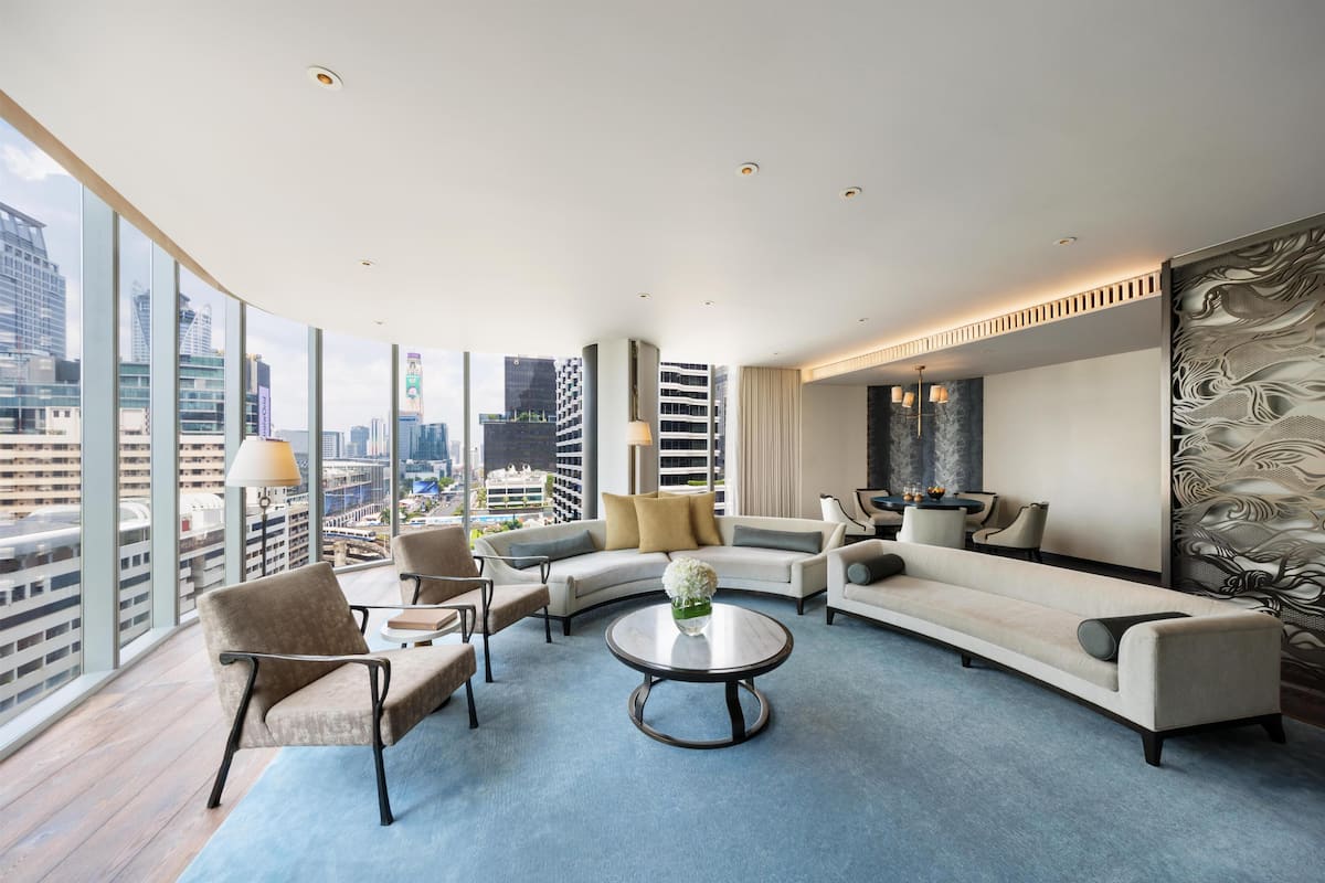 Een moderne hotelkamer met een gebogen bank, fauteuils, een ronde salontafel en een groot raam met uitzicht op de stad.