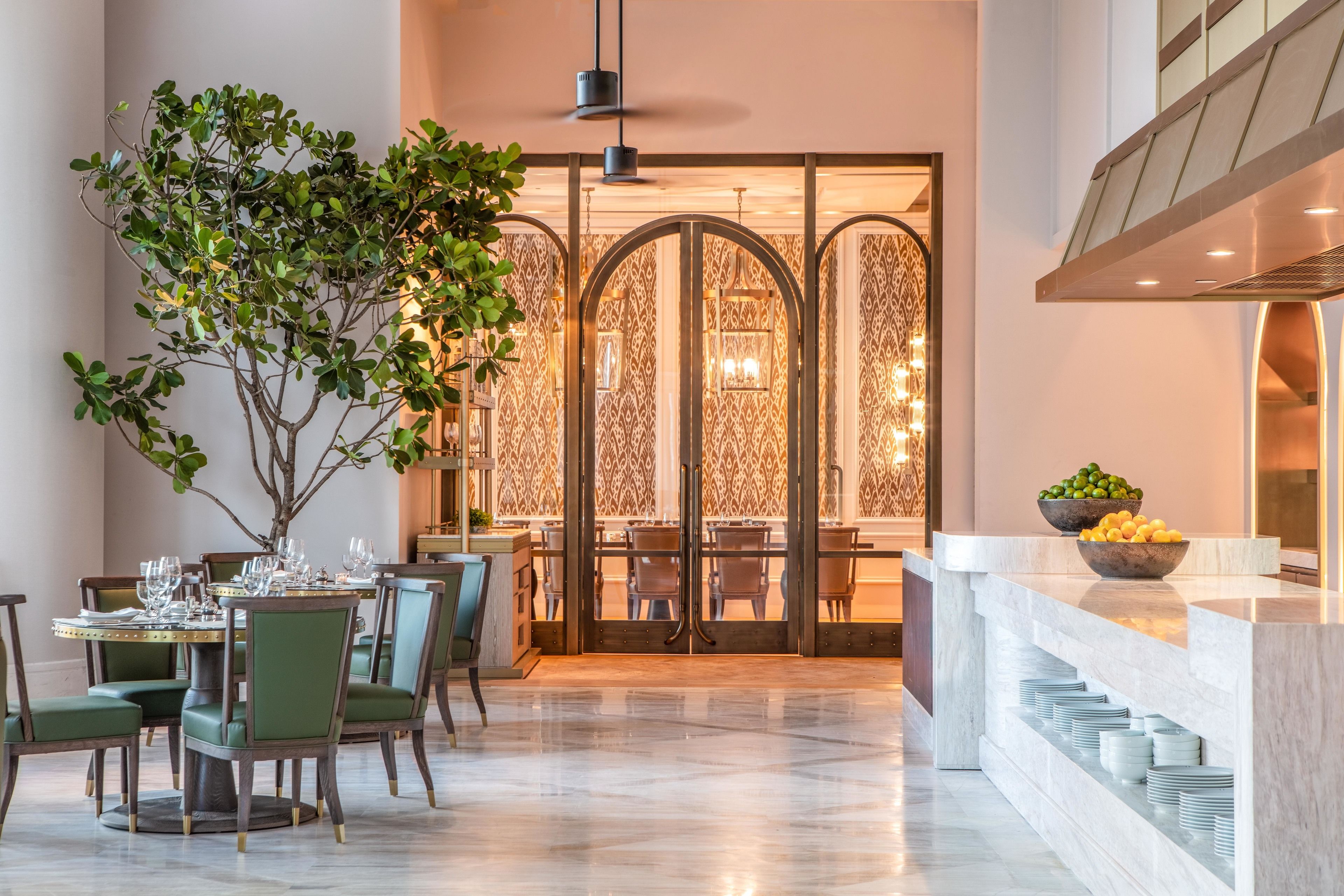 A dining area with a round table, chairs, and a potted tree. There is a buffet counter with plates and bowls of fruit.