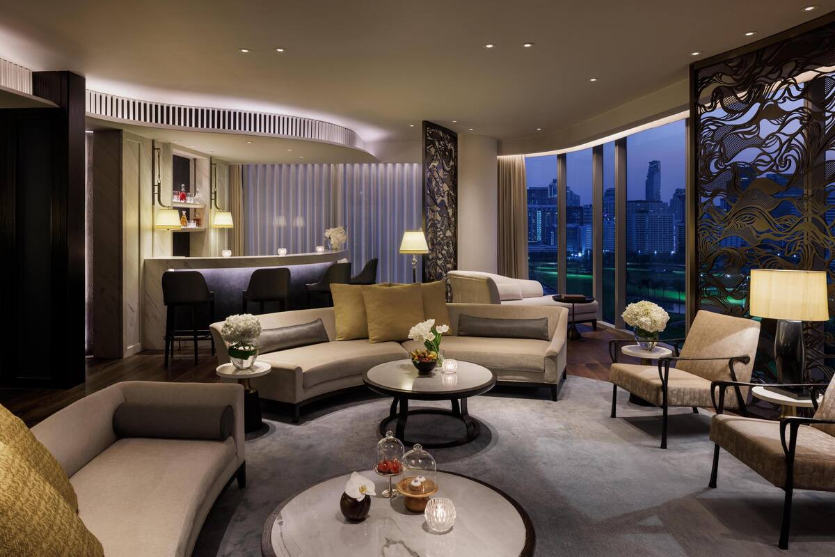 Een moderne hotelkamer met een gebogen zithoek, een ronde salontafel met decoratieve objecten, uitzicht op de stads skyline door grote ramen, en een decoratieve wand met gedetailleerde patronen.