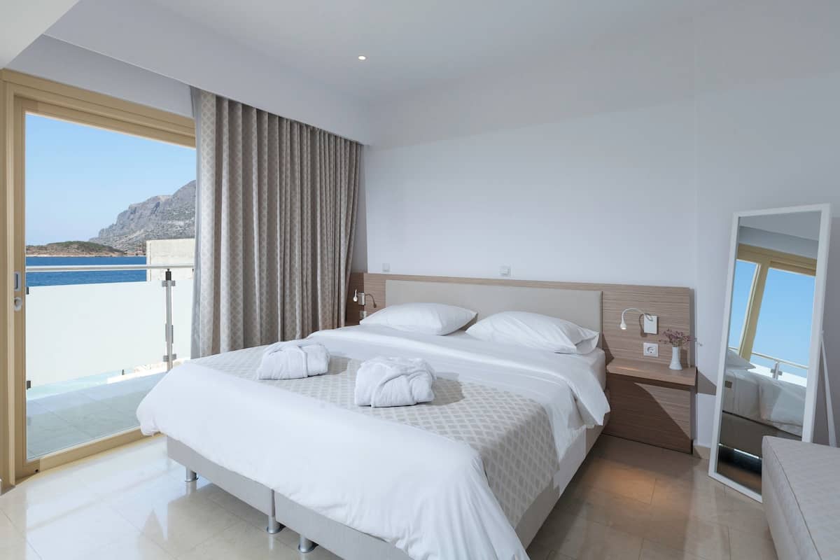 A hotel room with a large bed, a bedside table with a lamp, a mirror, and a view of the sea.