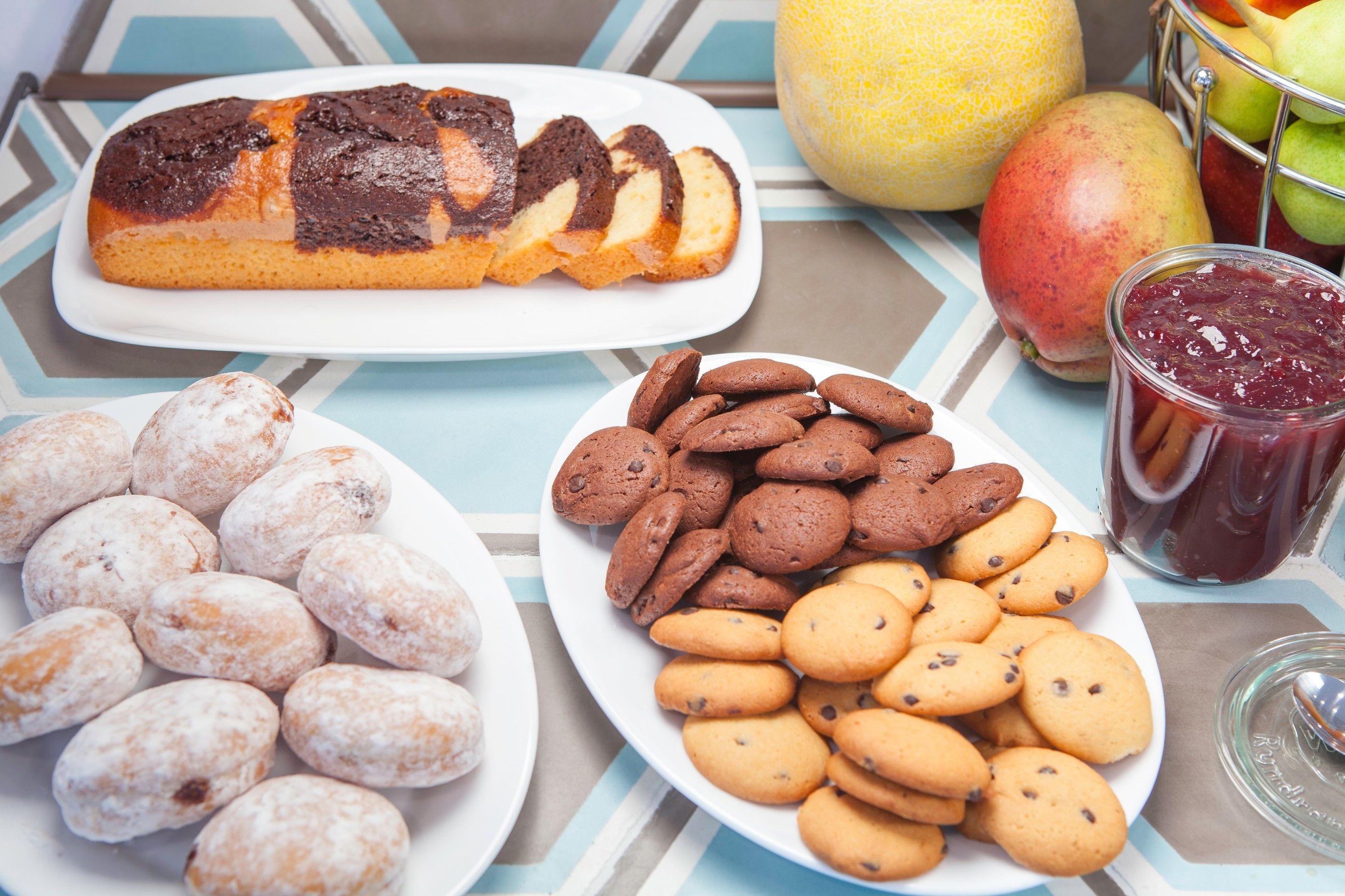 Una mesa con pan, platos de galletas, un frasco de mermelada y un melón.