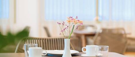 Una mesa de comedor con tazas y platillos blancos, un florero blanco con flores y un vaso transparente.