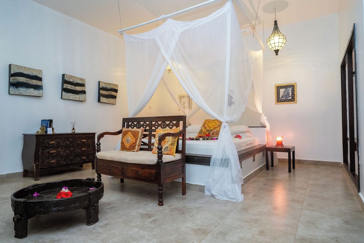 A bedroom with a bed under a canopy, a wooden sofa, a nightstand, a small table with a candle, and artwork on the walls.