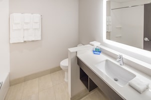 Un baño moderno con un lavamanos, espejo y retrete blancos. Toallas blancas cuelgan en la pared y hay una pequeña botella azul sobre el mostrador.