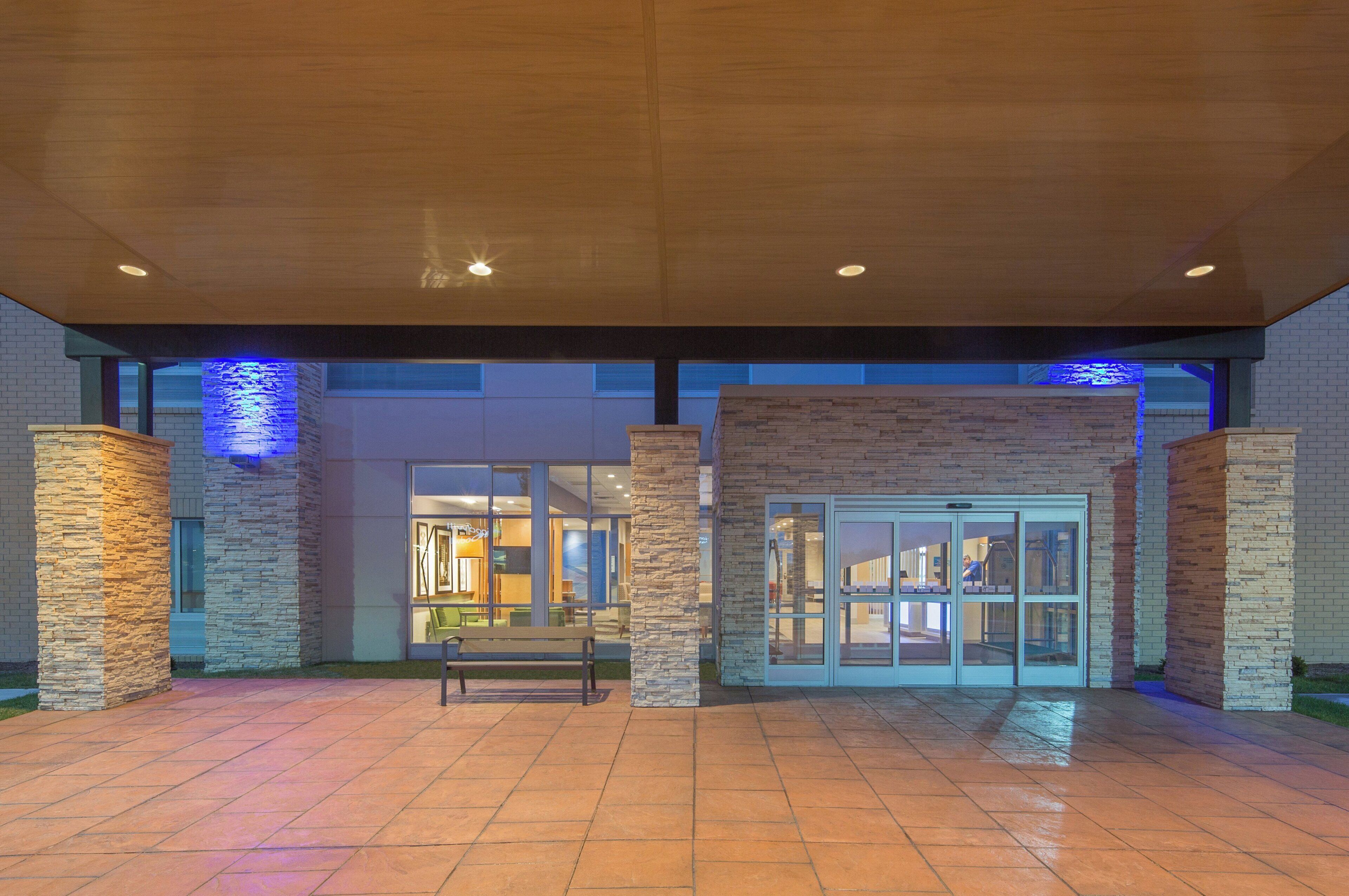 A covered outdoor area with stone pillars, a bench, and a building entrance with glass doors.