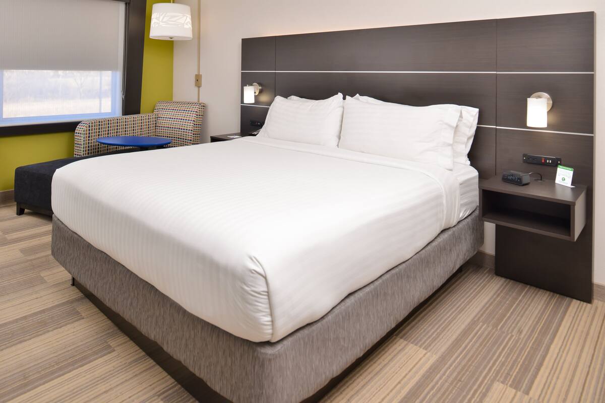 A modern hotel room with a large bed, a small table, a sofa, and a nightstand with a lamp.