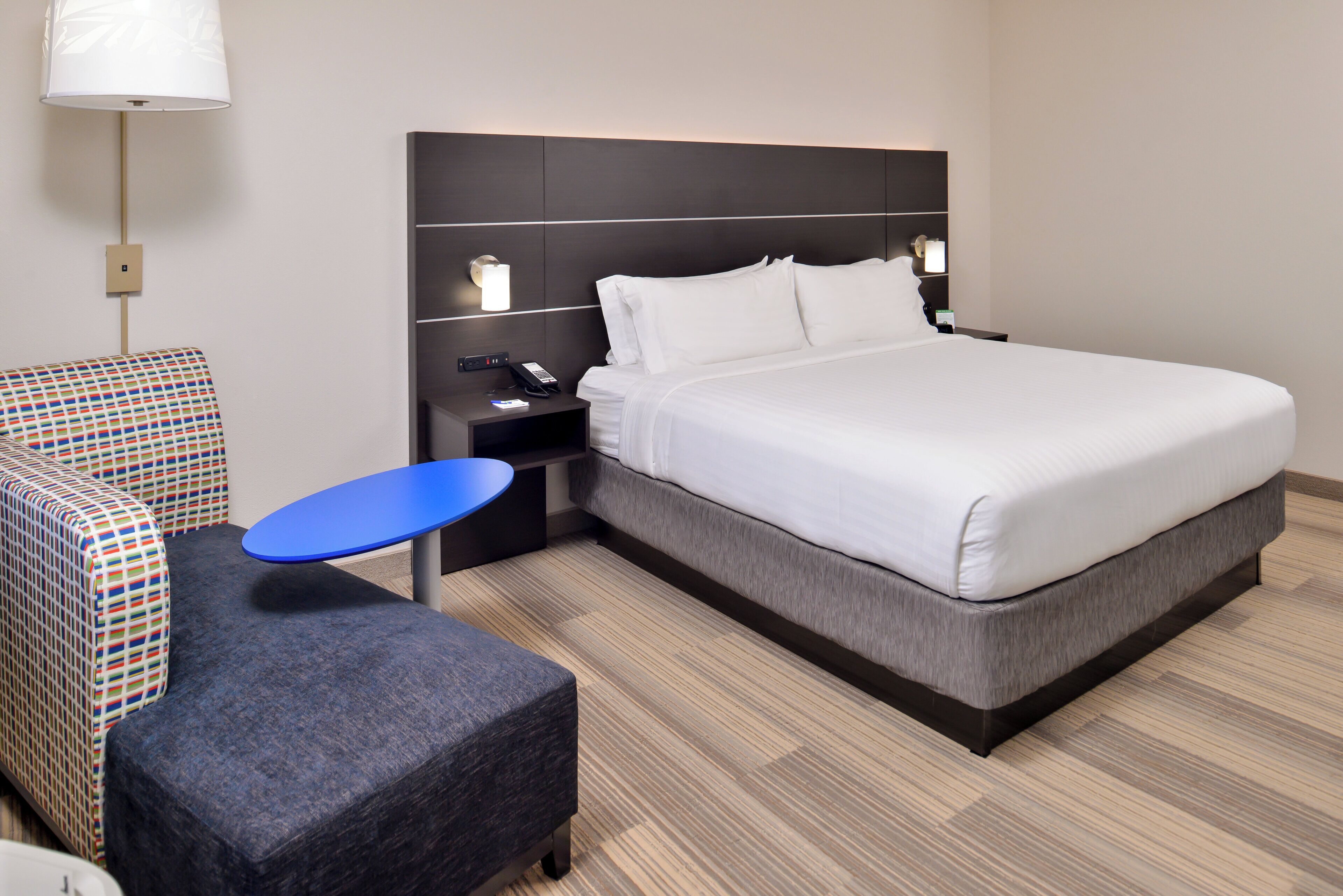 A hotel room with a large bed, a small table, a chair, a nightstand, and a lamp.