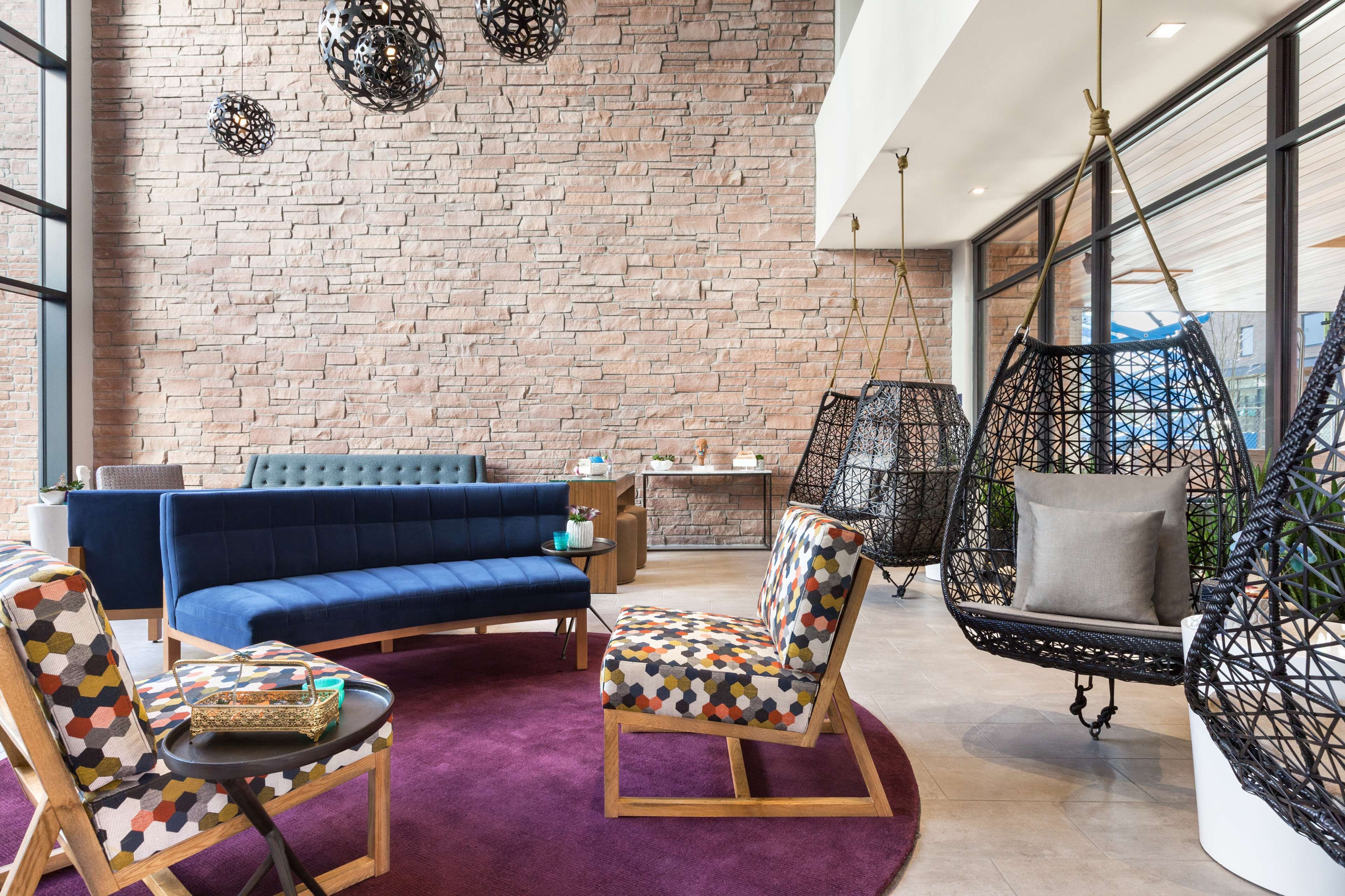A modern lounge area with a brick wall, blue sofa, patterned chairs, and a hanging chair.
