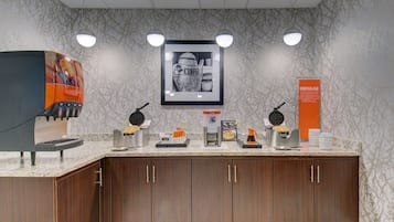 A coffee station with multiple coffee makers and a framed coffee-themed artwork on the wall.