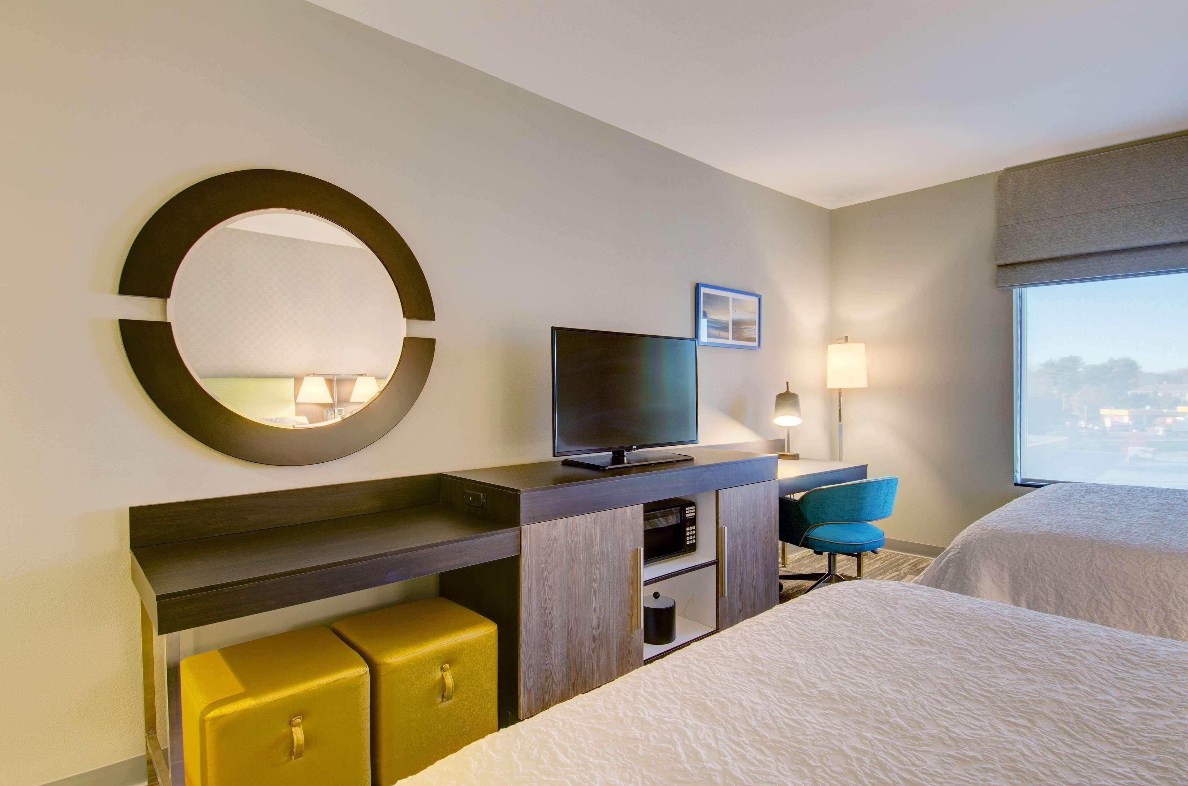 A hotel room with a bed, a TV, a desk with a lamp, a chair, and a large window.