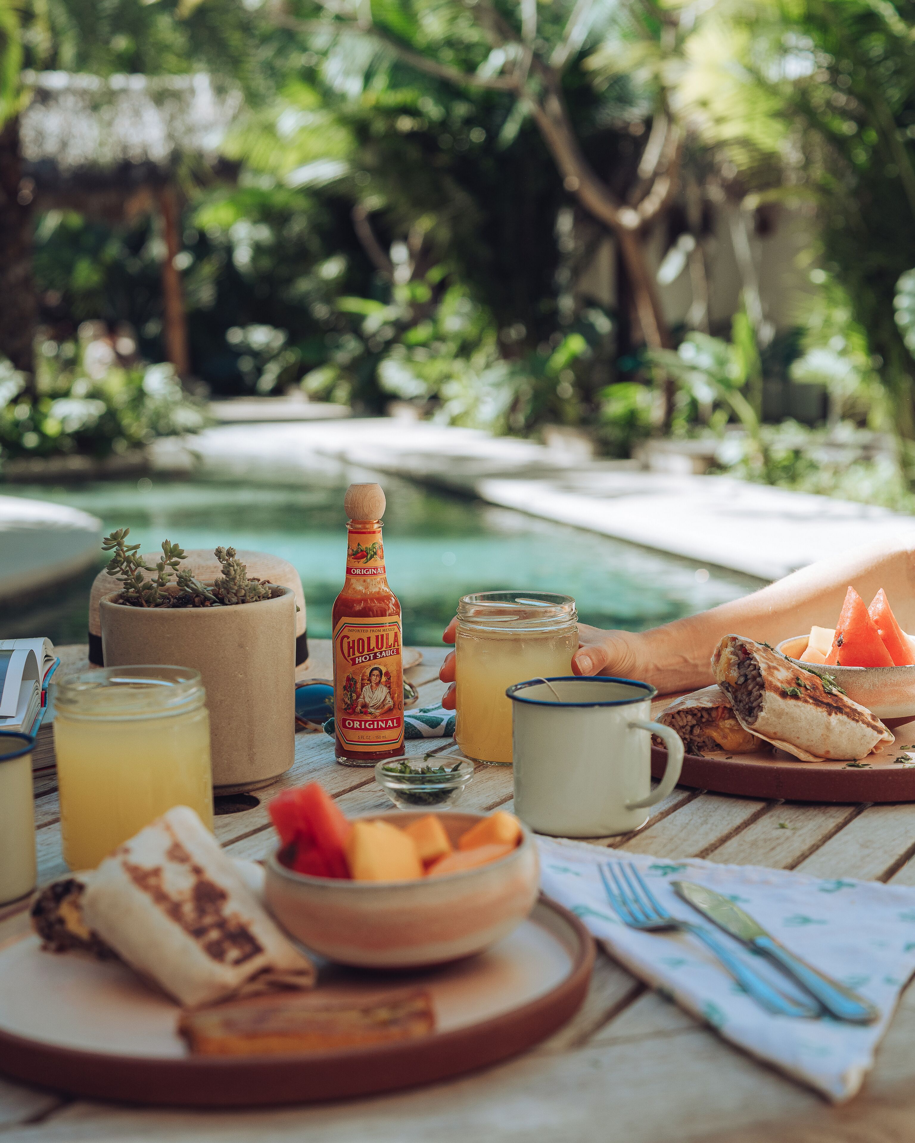 Una mesa de comedor con comida y bebidas, una botella de salsa picante, y una piscina al fondo.