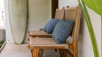 A wooden chair with woven seats and cushions, a surfboard leaning against a wall, and a large green plant in the foreground.