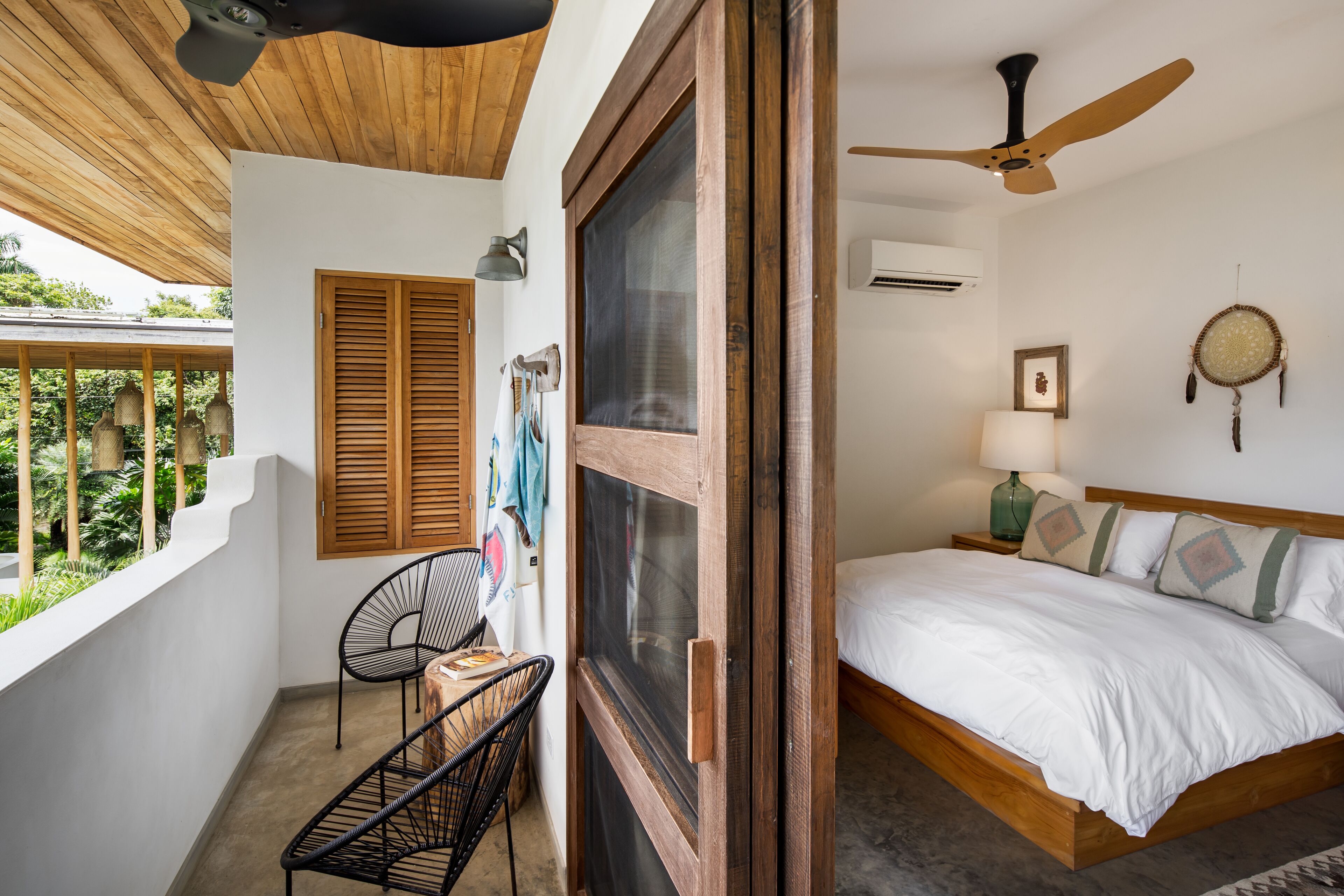 A bedroom with a bed, a ceiling fan, a window with wooden shutters, and a balcony with a chair and table.